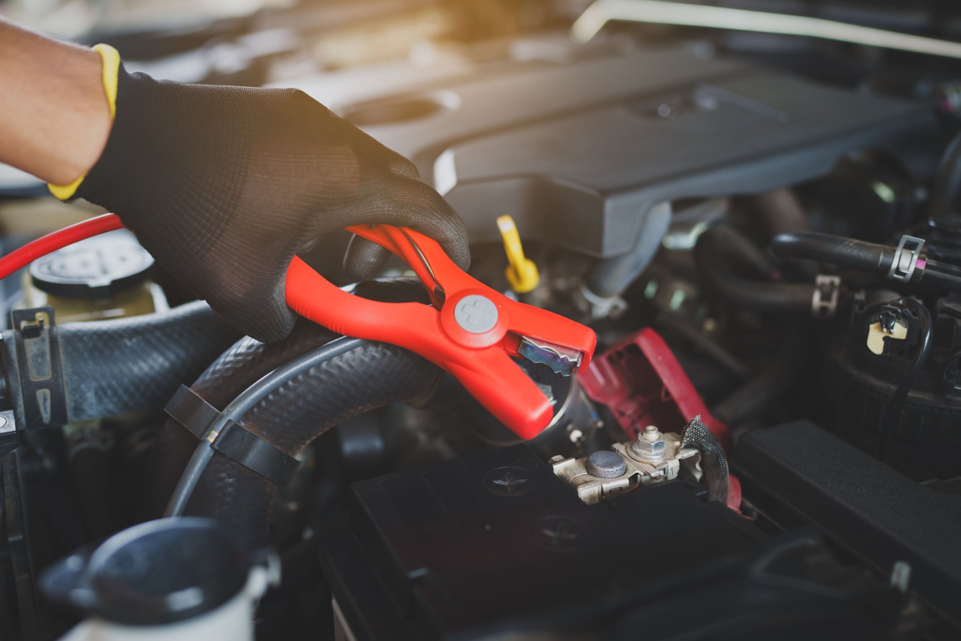 Hands in black gloves are carefully attaching red jumper cables to a car battery terminal, ready to jump-start a dead battery.